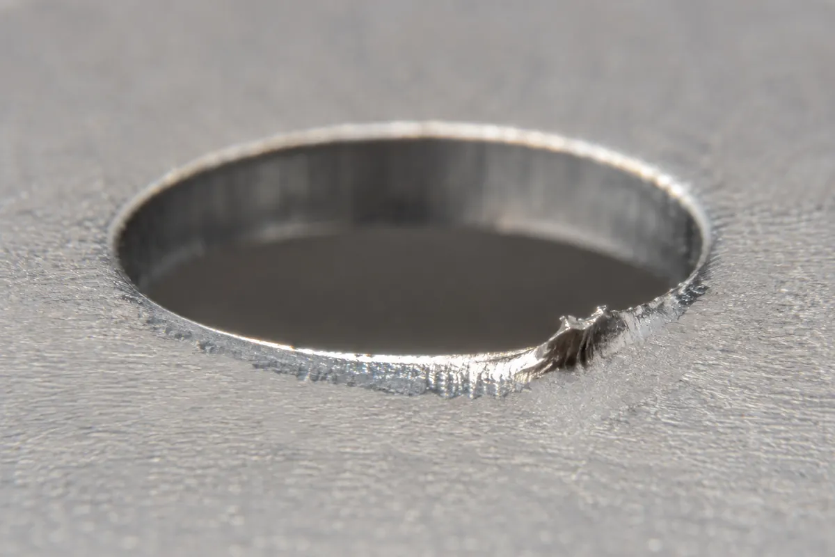 macro of a punched mounting hole edge and lifted burr/slug witness on sheet metal, realistic scale.